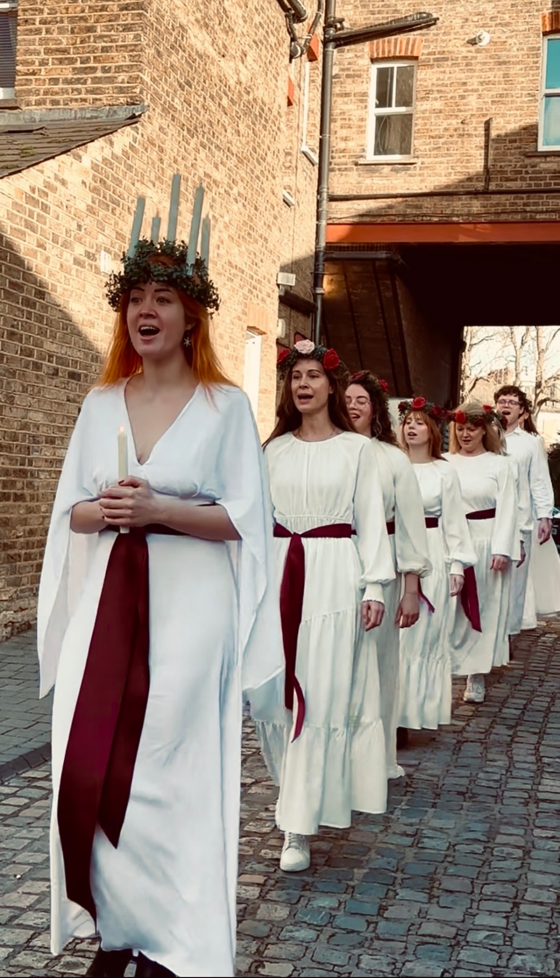 Lucia procession in white robes with red sashes walking through a London street