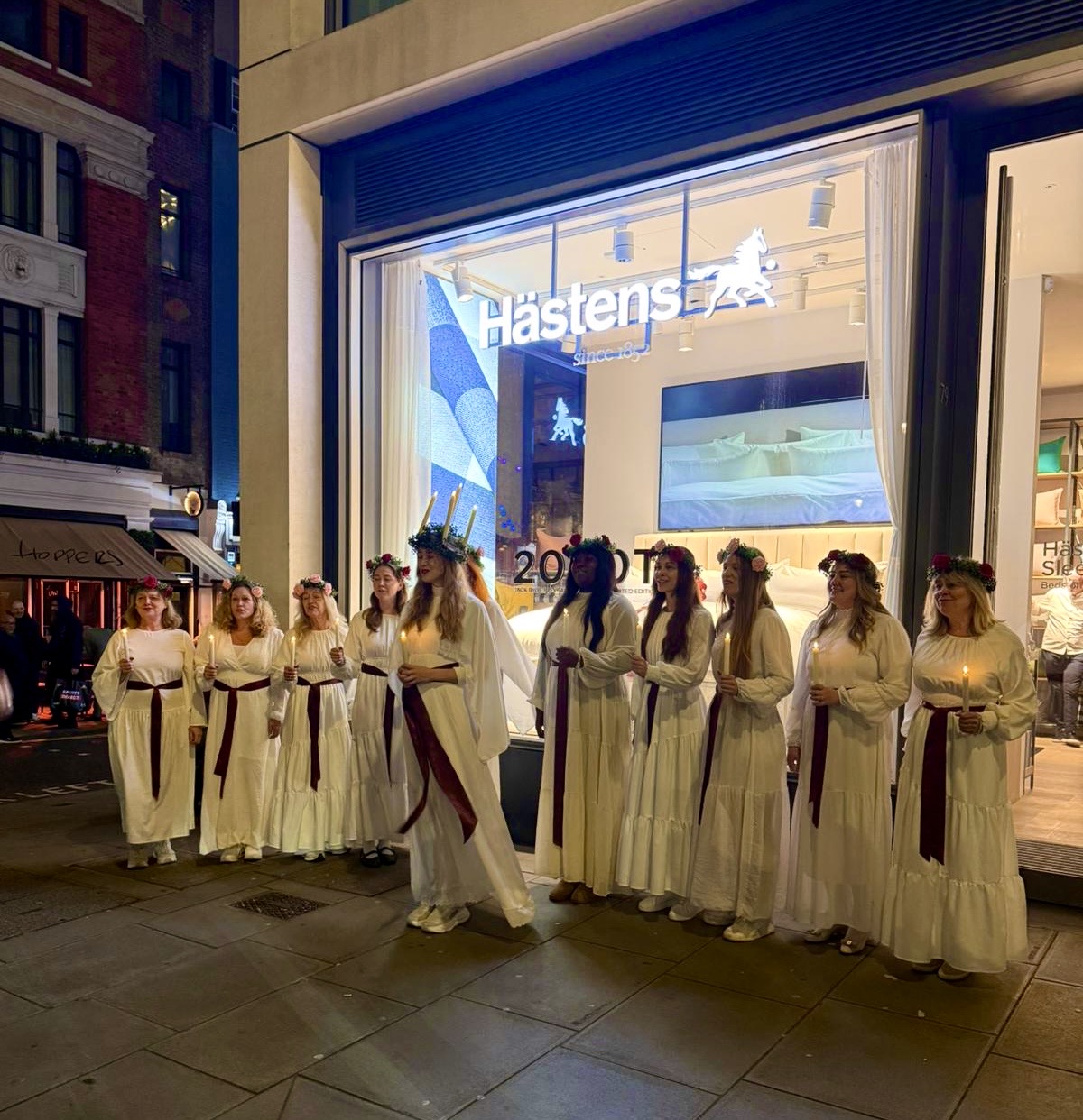 LondonSwedes Choir in white Lucia robes outside the Hästens store at night