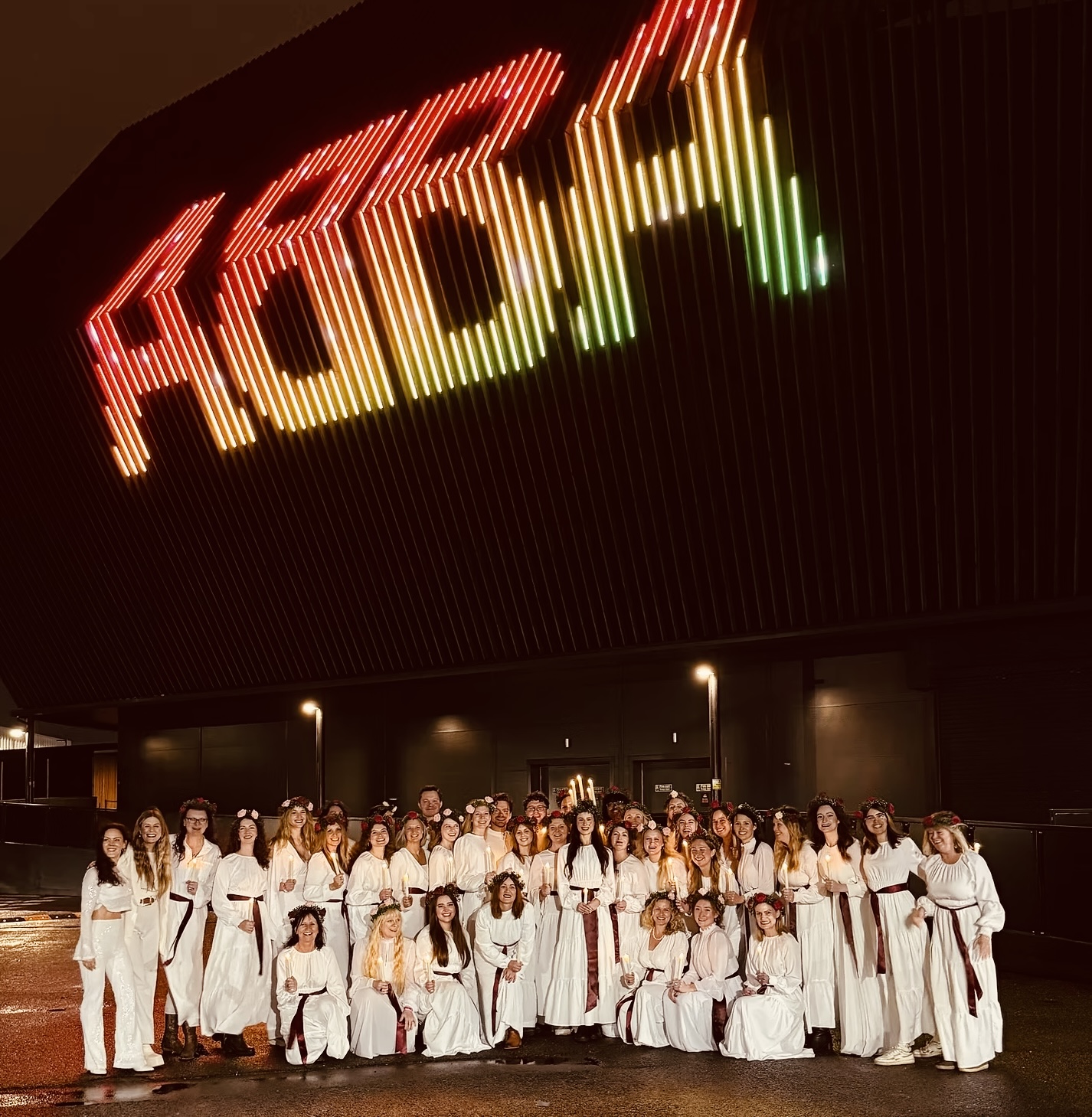 Full choir in white Lucia robes outside the ABBA Arena at night